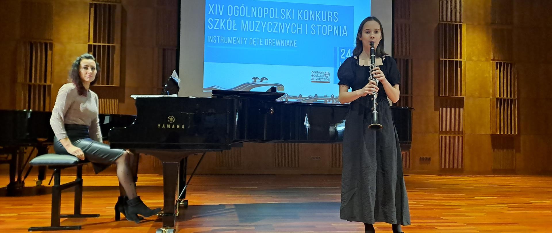 Na sali koncertowej stoi klarnecistka z instrumentem. Na drugim planie widać fortepian i siedzącą przy nim akompaniatorkę. W tel tablica świetlne z informacjami o konkursie.