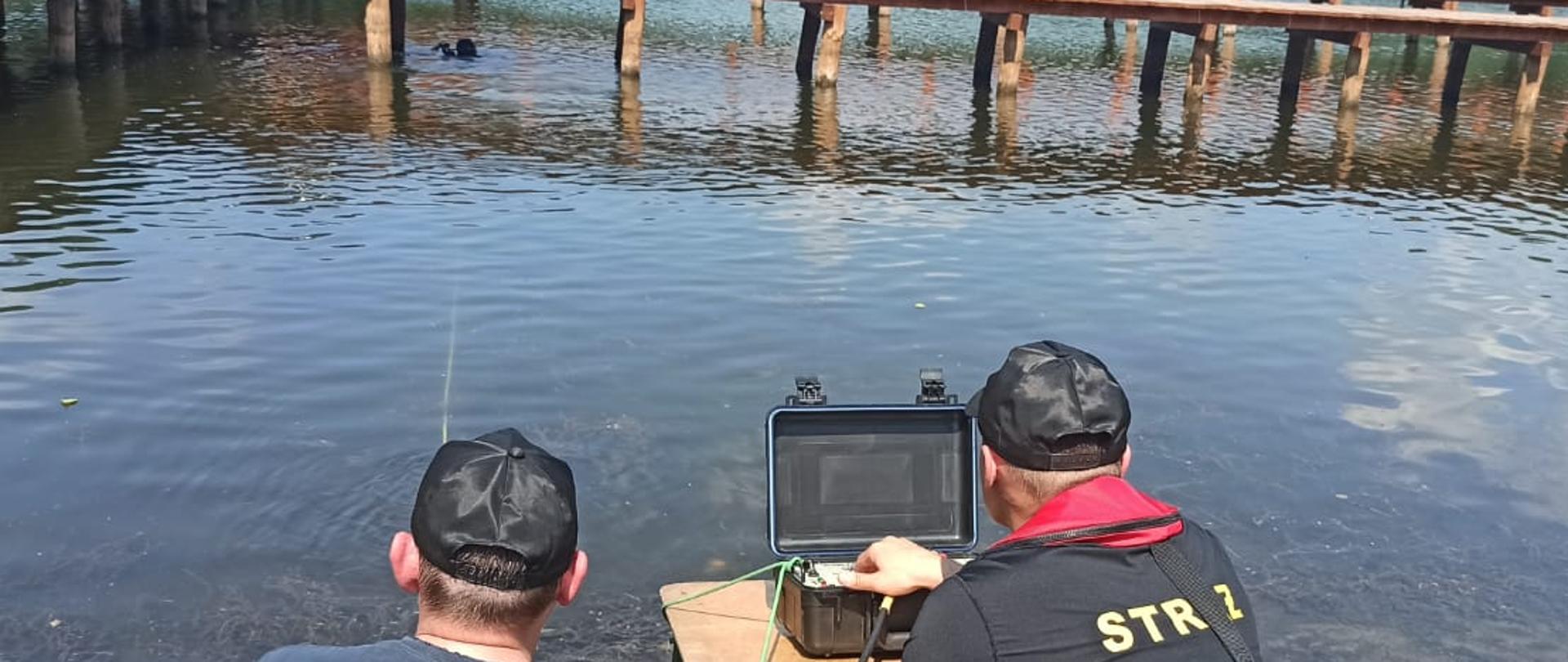 Zdjęcie przedstawia Jezioro Bukowiec oraz dwóch strażaków nadzorujących pracę nurka. W tle widać pomost.