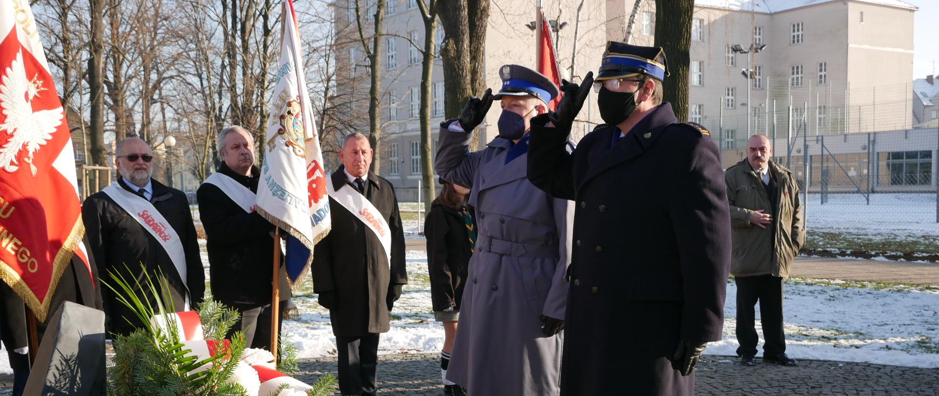 Strażak Państwowej Straży Pożarnej oraz policjiant oddają honory przez salutowanie przed pomnikiem solidarności. W tle poczty sztandarowe. Po lewej stronie na dole widoczne wieńce