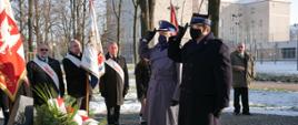 Strażak Państwowej Straży Pożarnej oraz policjiant oddają honory przez salutowanie przed pomnikiem solidarności. W tle poczty sztandarowe. Po lewej stronie na dole widoczne wieńce
