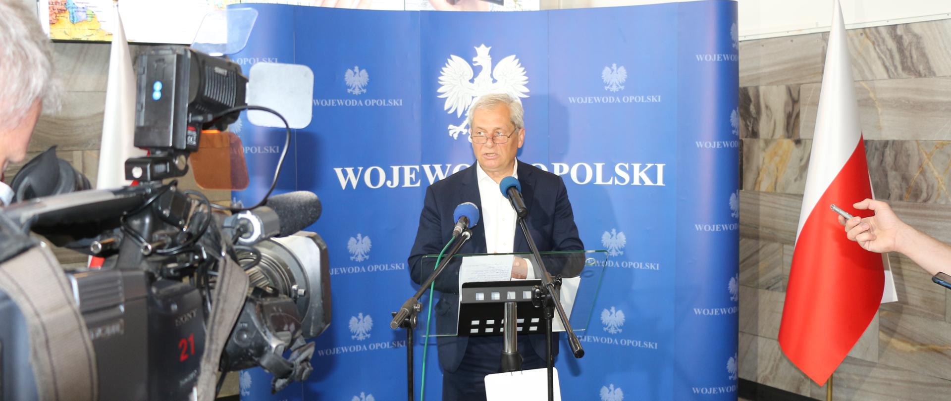 Na zdjęciu wojewoda opolski, przemawiający do zgromadzonych na spotkaniu. W tle granatowa ścianka z napisem Wojewoda Opolski. Przed wojewodą stoi szklana mównica i 2 mikrofonu na statywach. Za wojewodą w dalszej perspektywie dwie biało-czerwone flagi ulokowane naprzeciw siebie. W tle widać duży ekran monitora multimedialnego.
Na pierwszym planie z przodu widać kamerę telewizyjną.