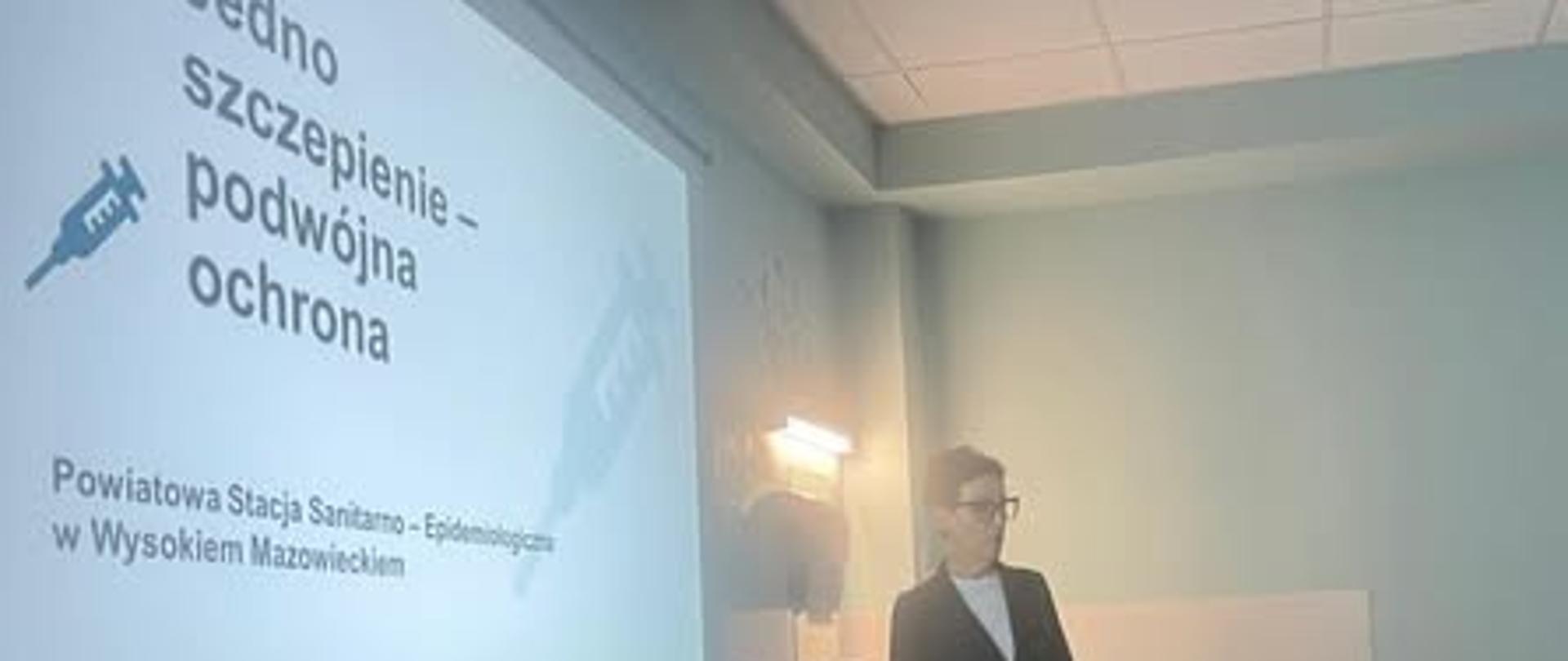 Fotografia przedstawiająca panią pracownik PSSE prowadzącą szkolenie. Za jej plecami wyświetlony slajd prezentacji z napisami: "Jedno szczepienie - podwójna ochrona.