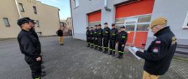 Komendant Powiatowy Państwowej Straży Pożarnej starszy brygadier Maciej Suchomski ubrany w mundur służbowy stoi naprzeciw Zastępcy Jednostki Ratowniczo Gaśniczej młodszego brygadiera Roberta Lissa trzymając w rękach otwarte pismo rozkazu. Po jego lewej i prawej stronie w szeregu ubrani w mundury koszarowe stoją na baczność naprzeciw siebie strażacy dwóch zmian służbowych.