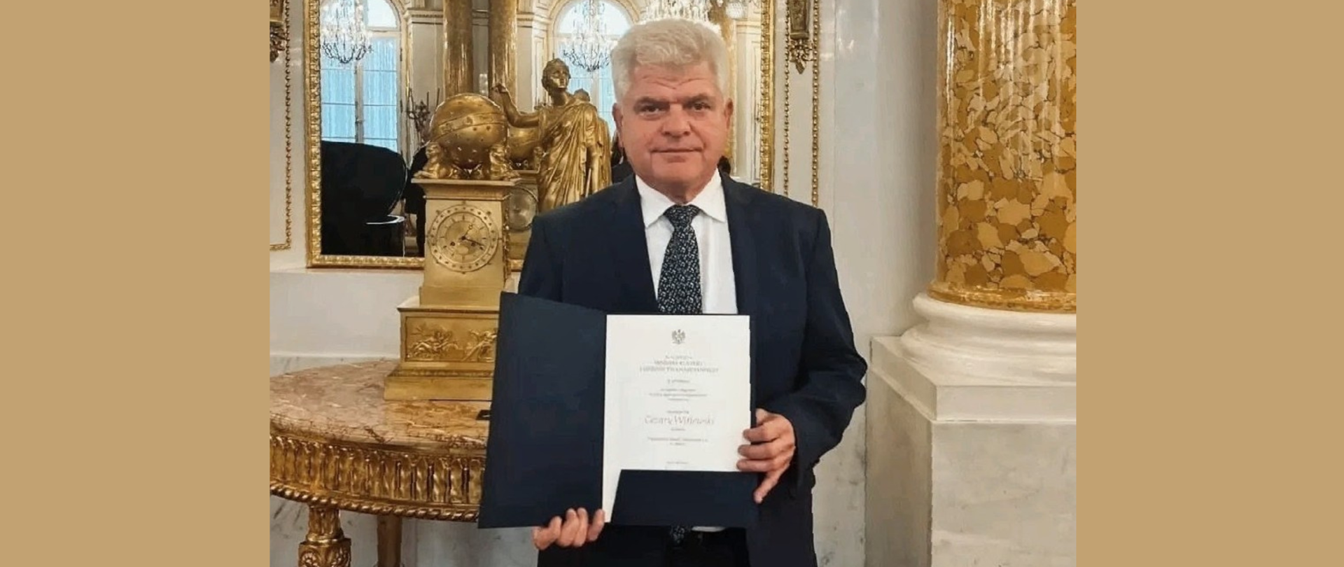 mężczyzna w garniturze trzyma otwarty oficjalny dokument w bogato zdobionej, złotej sali pałacowej.