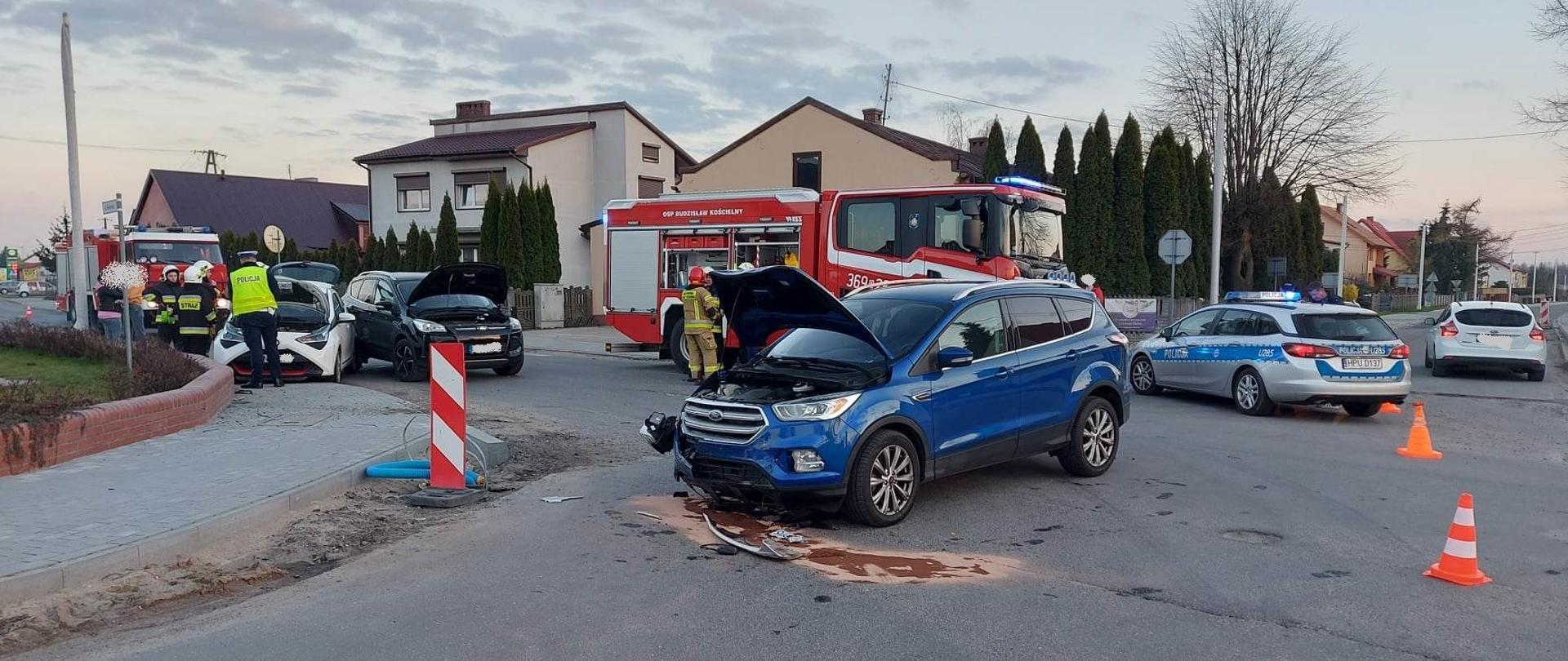 Zdjęcie przedstawia wypadek z udziałem trzech samochodów osobowych na skrzyżowaniu. Samochód koloru niebieskiego stoi przodem z otwartą maską. Kolejne dwa samochody widać z lewej strony (kolor biały i czarny) z otwartymi maskami. Na zdjęciu widać również strażaków, samochód strażacki i partol policji. Droga zablokowana pachołkami. 