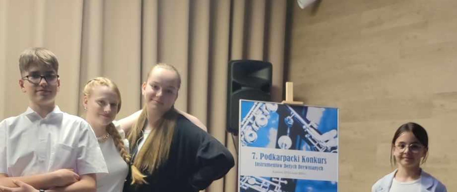 Grupa uśmiechniętych uczniów stoi w Sali koncertowej w PSM w Kamieniu. Wszyscy są ubrani w galowe stroje. W tle widnieje plakat konkursu. Ogólna kolorystyka czarno-beżowa.