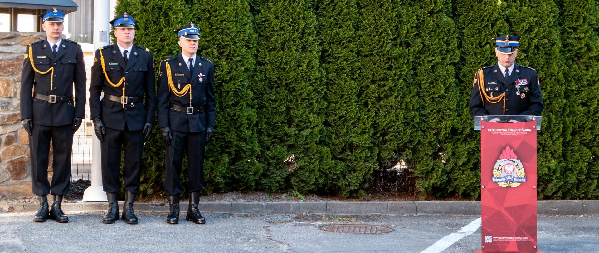 Zdjęcie przedstawia poczet flagowy i przemawiającego przy mównicy komendanta powiatowego z Ustrzyk Dolnych. Zdjęcie zostało wykonane na przednim placu Komendy Powiatowej PSP w Ustrzykach Dolnych.