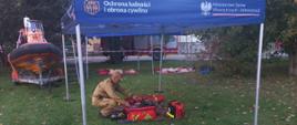 Widok na niebieski namiot wystawienniczy z białym napisem „Ochrona ludności i obrona cywilna” oraz logo Ministerstwa Spraw Wewnętrznych i Administracji. Pod namiotem na trawie strażak klęczy przy rozłożonym sprzęcie i torbach ratowniczych. Po lewej stronie w tle stoi pomarańczowa łódź ratownicza na przyczepie oraz wóz strażacki.