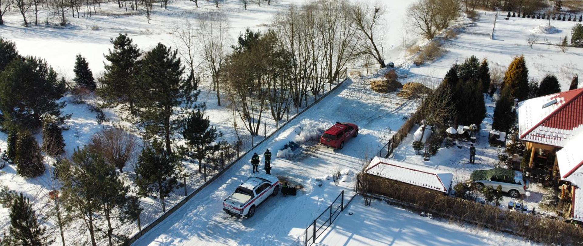 Zdjęcie przedstawia widok z drona - dwa samochody operacyjne Państwowej Straży Pożarnej ustawione na drodze. Po prawej stronie widać zabudowania prywatne natomiast po lewej widać zamarzniętą rzekę.
