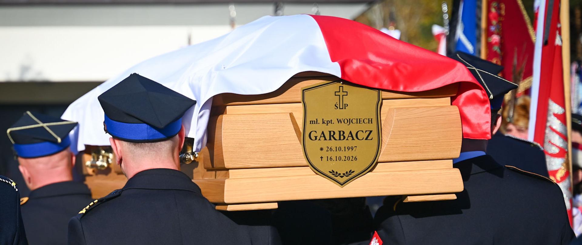 Strażacy Państwowej Straży Pożarnej w mundurach wyjściowych niosą na ramionach drewnianą trumnę przykrytą biało-czerwoną flagą Polski. Na trumnie widnieje tabliczka w kształcie tarczy z napisem: „mł. kpt. WOJCIECH GARBACZ * 26.10.1997 † 16.10.2026”.