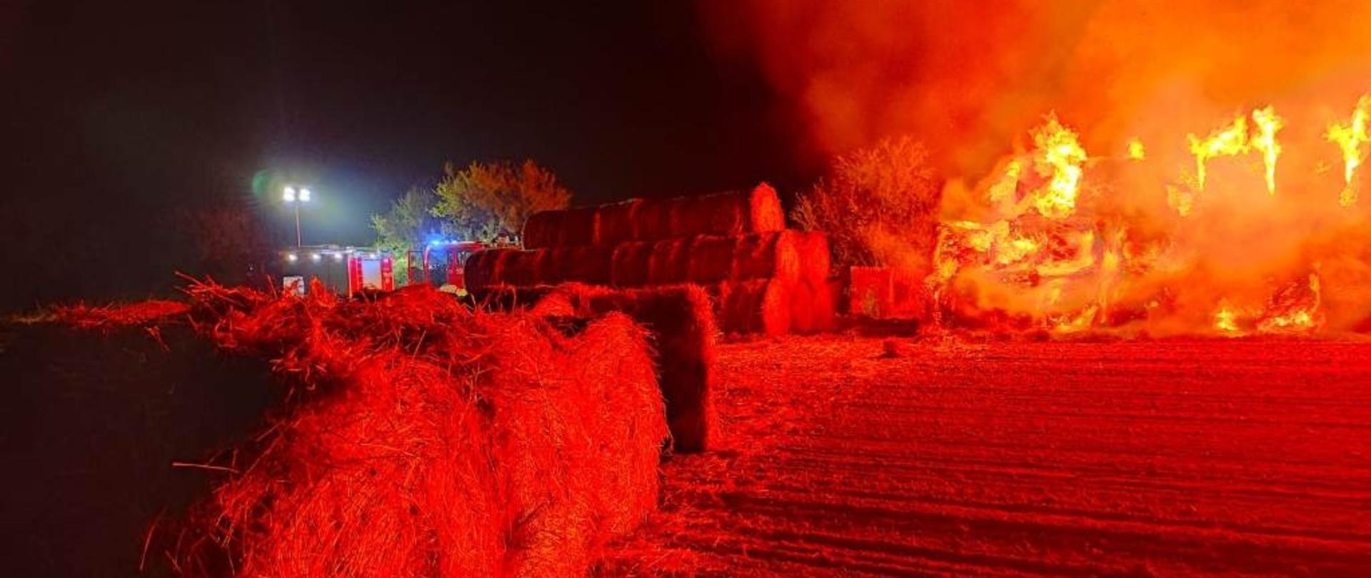 Płonący w całej objętości stóg słomy. Na bliższym planie widać odsunięte od płomieni nienaruszone baloty. W oddali stoją samochody strażackie.