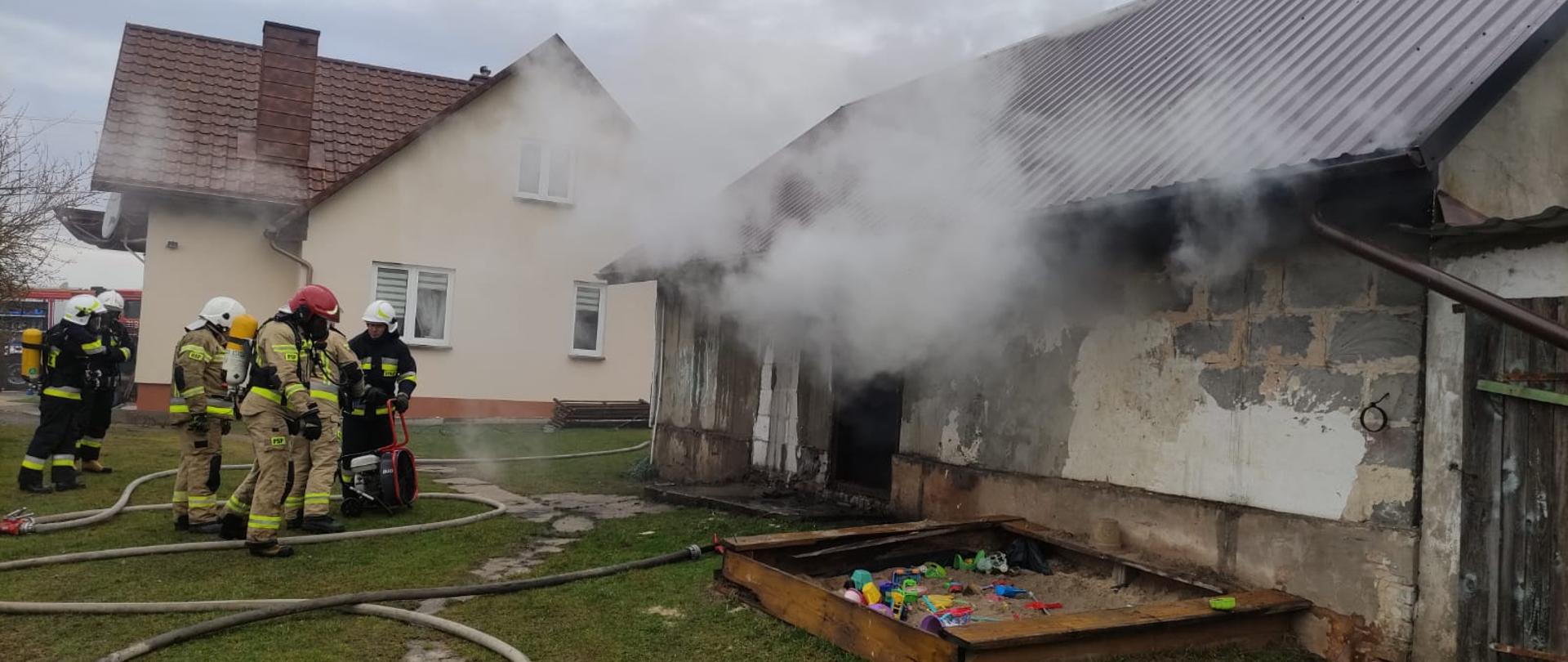 Zdjęcie przedstawia budynek gospodarczy (po prawej stronie) z którego otworów wydobywa się gesty dym. Grupa ratowników przygotowuje się do oddymiania pomieszczenia. Dwóch z nich stoi w aparatach dróg oddechowych. Na ziemi leżą rozwinięte węże gaśnicze. W tle widać budynek mieszkalny.