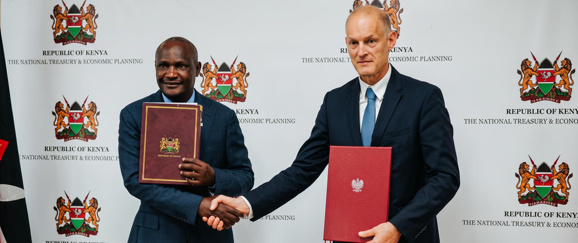 Dwie osoby w ciemnych garniturach stoją przed ścianką z herbem Republiki Kenii i napisem „The National Treasury & Economic Planning”. Osoby podają sobie dłonie i prezentują dokumenty w ozdobnych teczkach: bordowej z godłem Kenii oraz czerwonej z polskim orłem. Po lewej stronie kadru widoczny jest fragment flagi Kenii.
