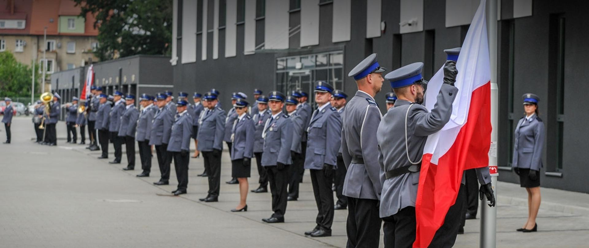 Poczet flagowy złożony z 3 policjantów wciąga biało-czerwoną flagę na maszt. w tle funkcjonariusze i funkcjonariuszki policji w galowych mundurach stoją w rzędach, zwróceni w stronę flagi.