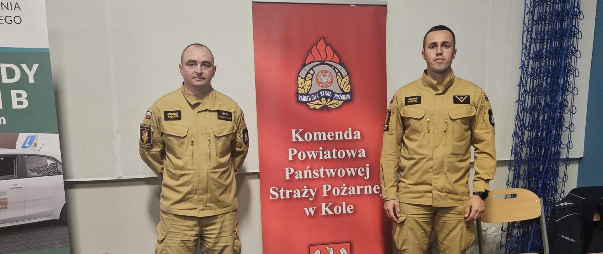 Na zdjęciu widać dwóch strażaków w jasnożółtych mundurach stojących po obu stronach czerwonego roll-upu z napisem „Komenda Powiatowa Państwowej Straży Pożarnej w Kole”. Przed nimi stoi stół, na którym leżą broszury informacyjne oraz elementy umundurowania, w tym hełm strażacki i kurtka ochronna. Scena stanowi stanowisko promocyjne podczas wydarzenia.