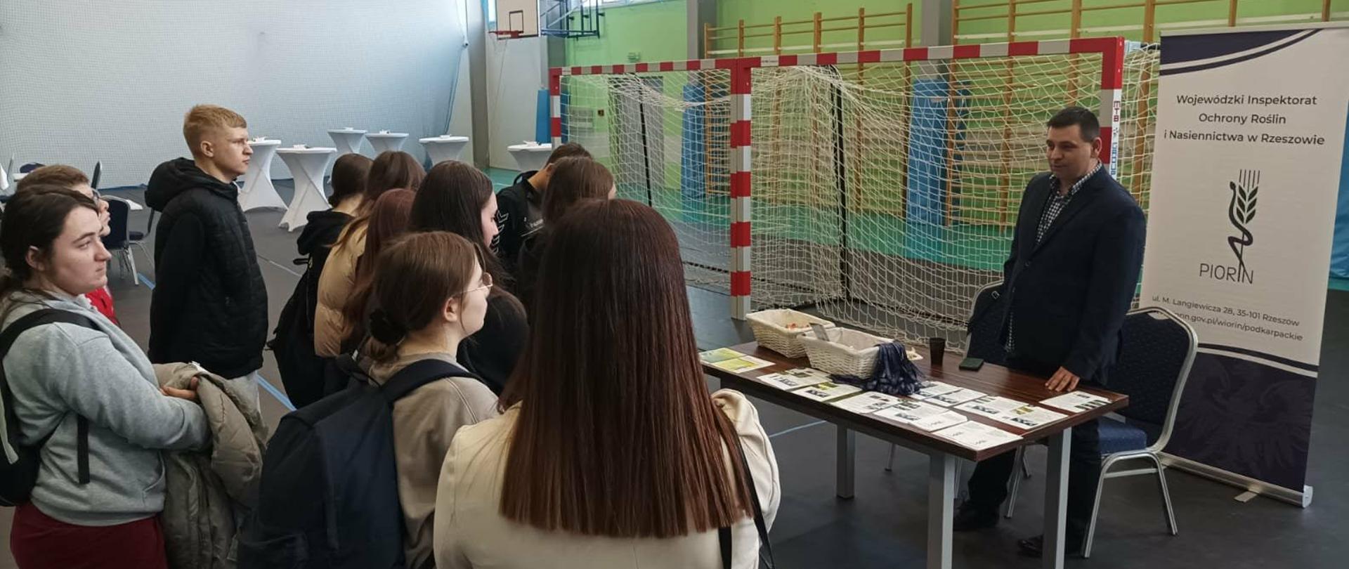 Prelegent w ciemnej marynarce stoi za stołem wystawowym i przemawia do grupy młodzieży zgromadzonej przed nim w dużej hali sportowej. Uczestnicy stoją blisko siebie, część z nich ma plecaki i kurtki. Na stole rozłożone są liczne materiały edukacyjne, a obok stoją koszyki z gadżetami. Za prelegentem ustawiony jest baner instytucji związanej z ochroną roślin i nasiennictwem. W tle widoczna jest bramka sportowa, kosz do koszykówki oraz wysokie okna wpuszczające dużo światła dziennego, co podkreśla przestronność hali.