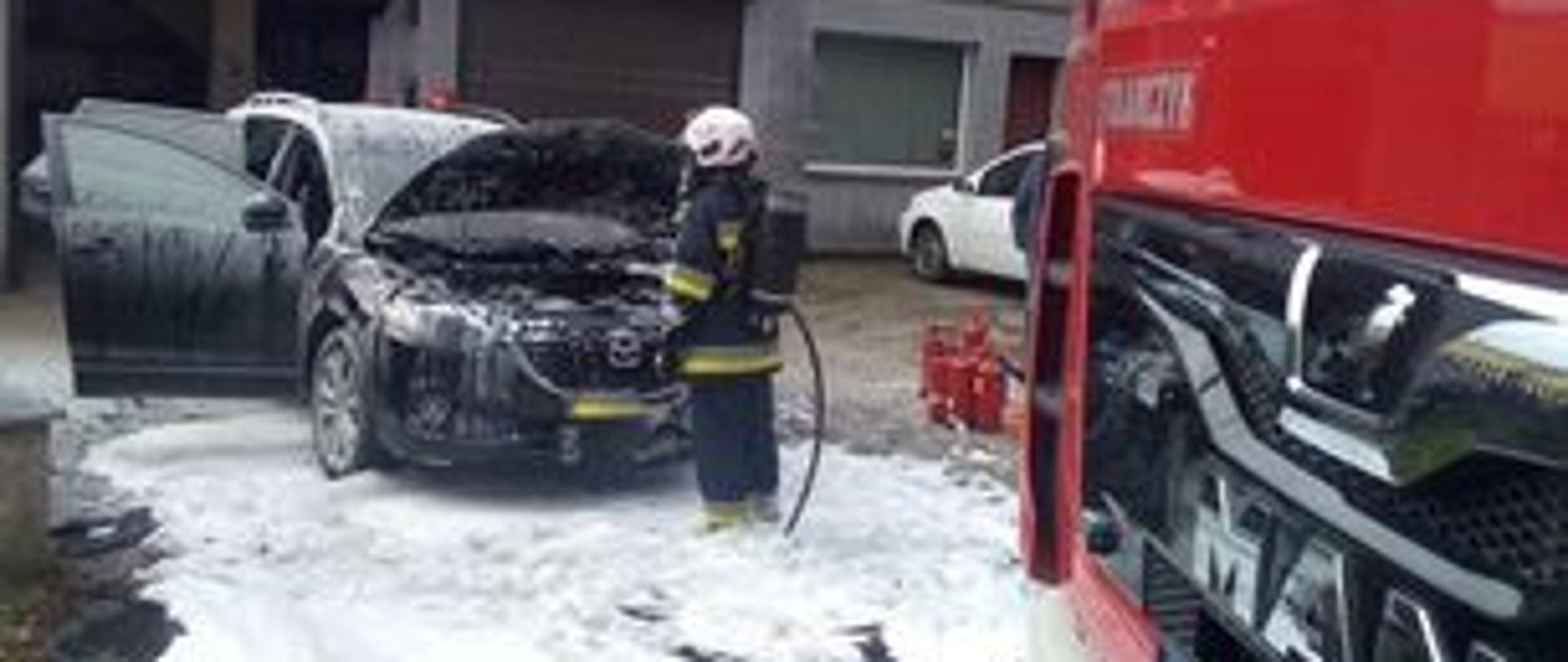 Zdjęcie przedstawia strażaków gaszących pożar samochodu osobowego.
W tle budynek mechanika samochodowego.

