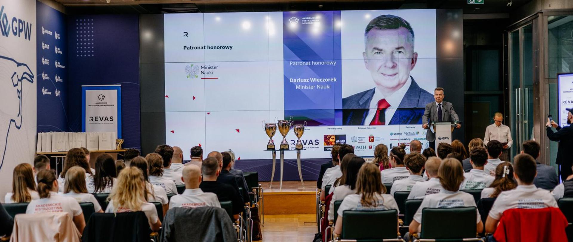 Widok od tyłu na salę, przy kilku stolikach siedzą grupki młodych ludzi, przed nimi z przodu sali minister Wieczorek stoi za mównicą i mówi do mikrofonu, za nim wielki ekran.