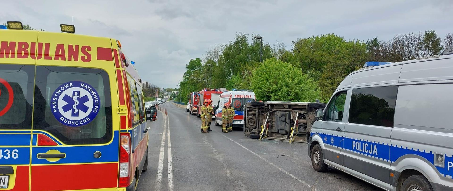 Służby ratownicze na drodze, na środku przewrócony ambulans transportowy zabezpieczony przy użyciu podpór stabilizujących.