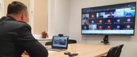 Sala konferencyjna, Komendant Powiatowy podczas prowadzenia wideokonferencji, w tle monitor wyświetlający uczestników spotkania on-line. 