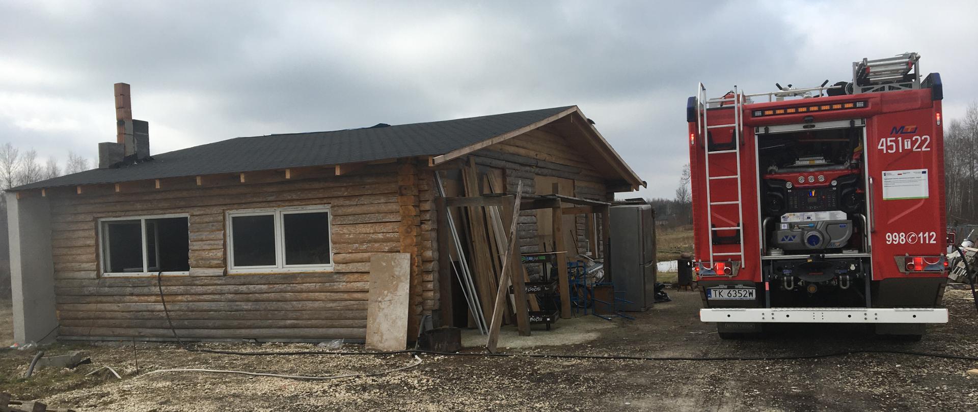 Na zdjęciu widać drewniany budynek gospodarczy, w którym doszło do pożaru trocin. Obok budynku stoi samochód gaśniczo-ratowniczy.