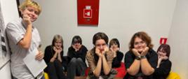 Grupa siedmiu młodych osób pozuje do zdjęcia w sali, siedząc przy stole i na kolorowych siedziskach. Na stole leżą zeszyty, przybory szkolne i, co sugeruje wspólną pracę lub spotkanie edukacyjne.