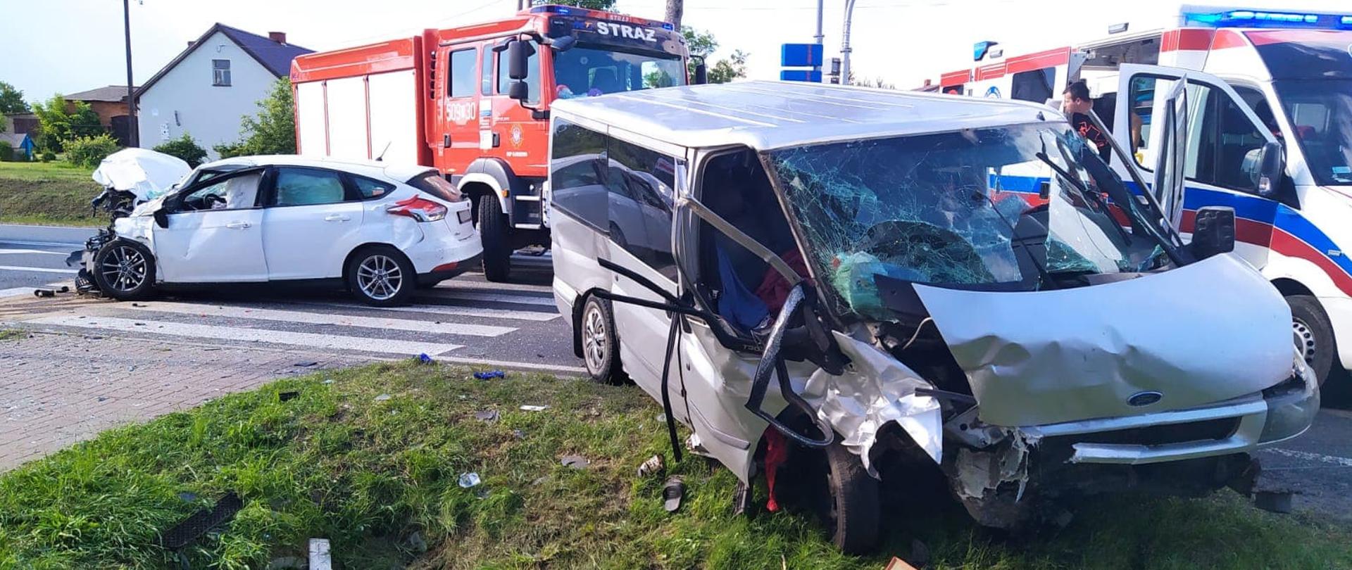 Dwa uszkodzone samochody Bus oraz osobowy stoją na drodze. W tle znajduje się samochód strażacki oraz karetka.