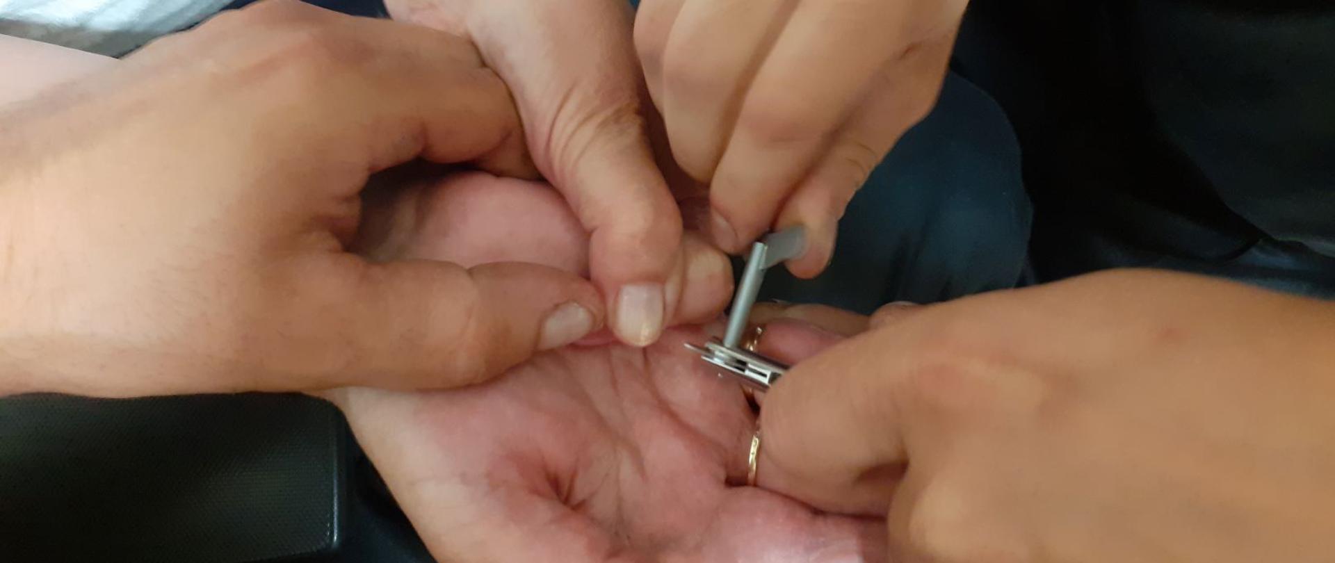 Zdjęcie przedstawia dłonie strażaków i poszkodowanej. Trwa przecinanie obrączki ręcznym urządzeniem 