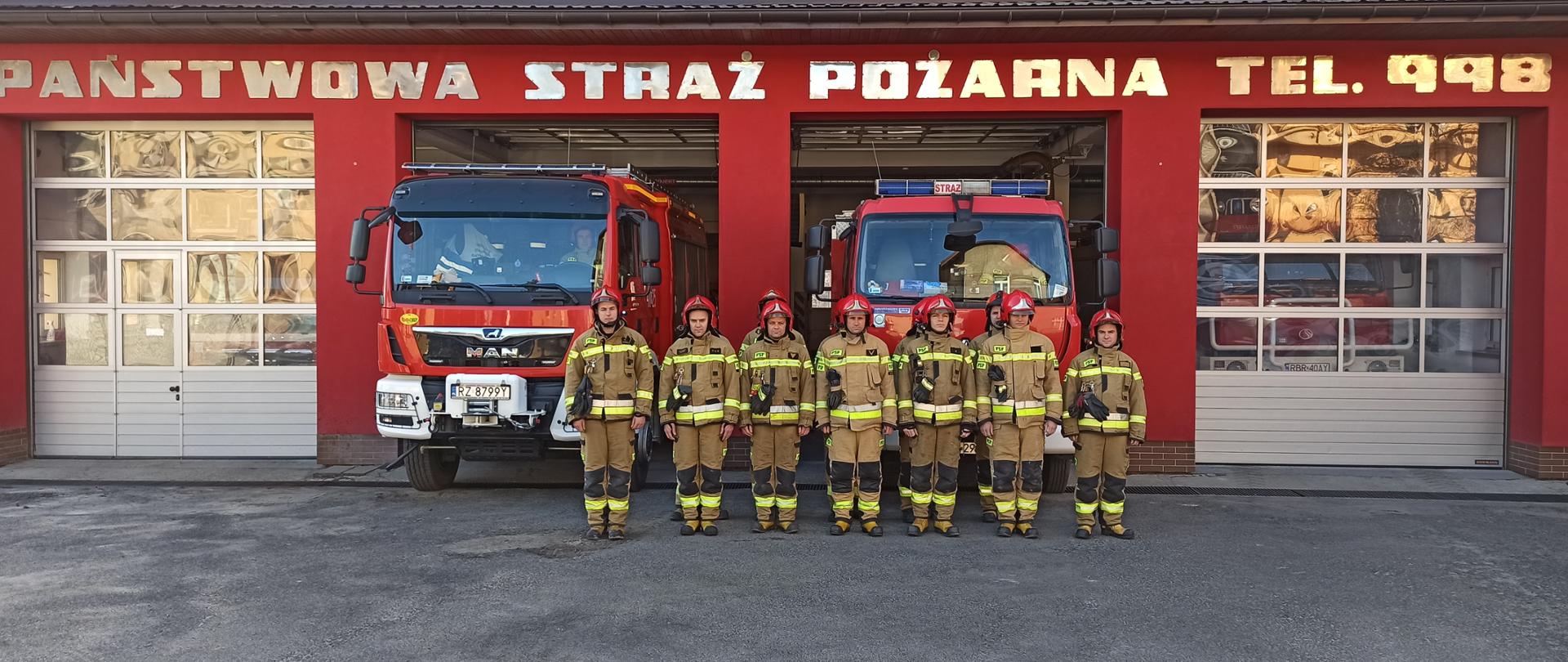 Uroczysta zbiórka i minuta ciszy w geście solidarności ze strażakami z Ukrainy. Na fotografii wykonanej w pogodny dzień widać strażaków na uroczystej zbiórce przed budynkiem strażnicy. W tle stoją samochody pożarnicze w bramach wyjazdowych budynku.