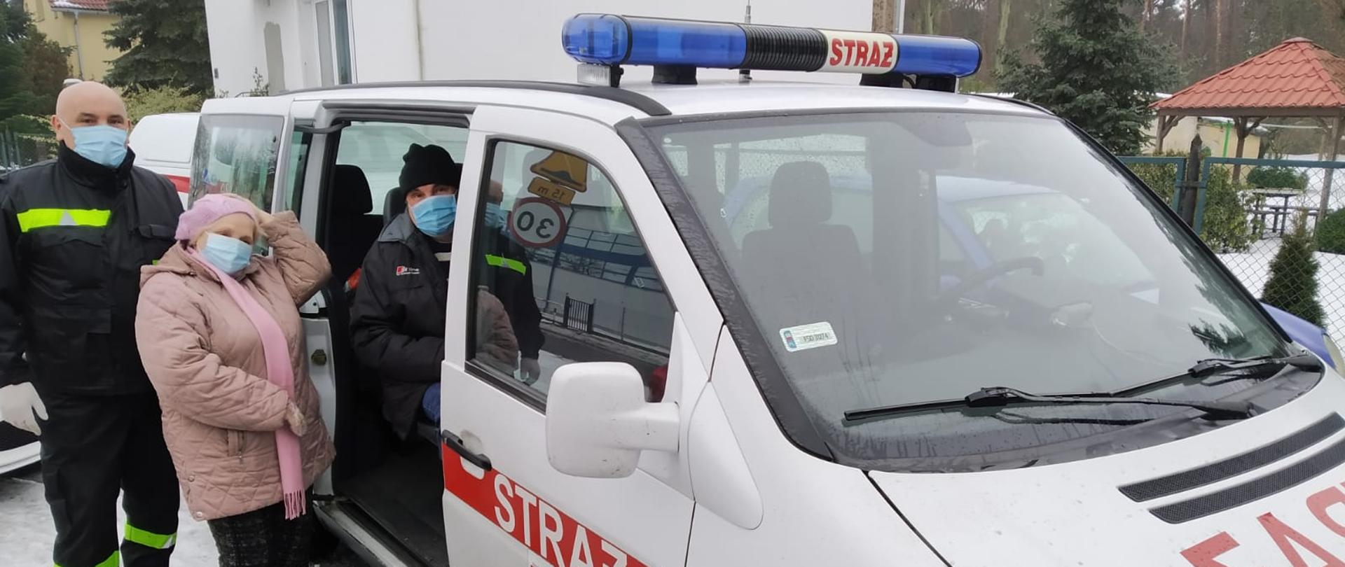 Fotografia przedstawia biały samochód strażacki typu van z otwartymi bocznymi przesuwnymi drzwiami. Ujęcie wzdłuż lewego boku pojazdu. W pojeździe na fotelu przy otwartych bocznych drzwiach siedzi mężczyzna w maseczce, obok niego na zewnątrz pojazdu stoi kobieta w maseczce i dalej obok kobiety strażak w umundurowaniu koszarowym także w maseczce. Pora zimowa, na ziemi leży na wpół roztopiony śnieg, w tle zabudowania mieszkalne.