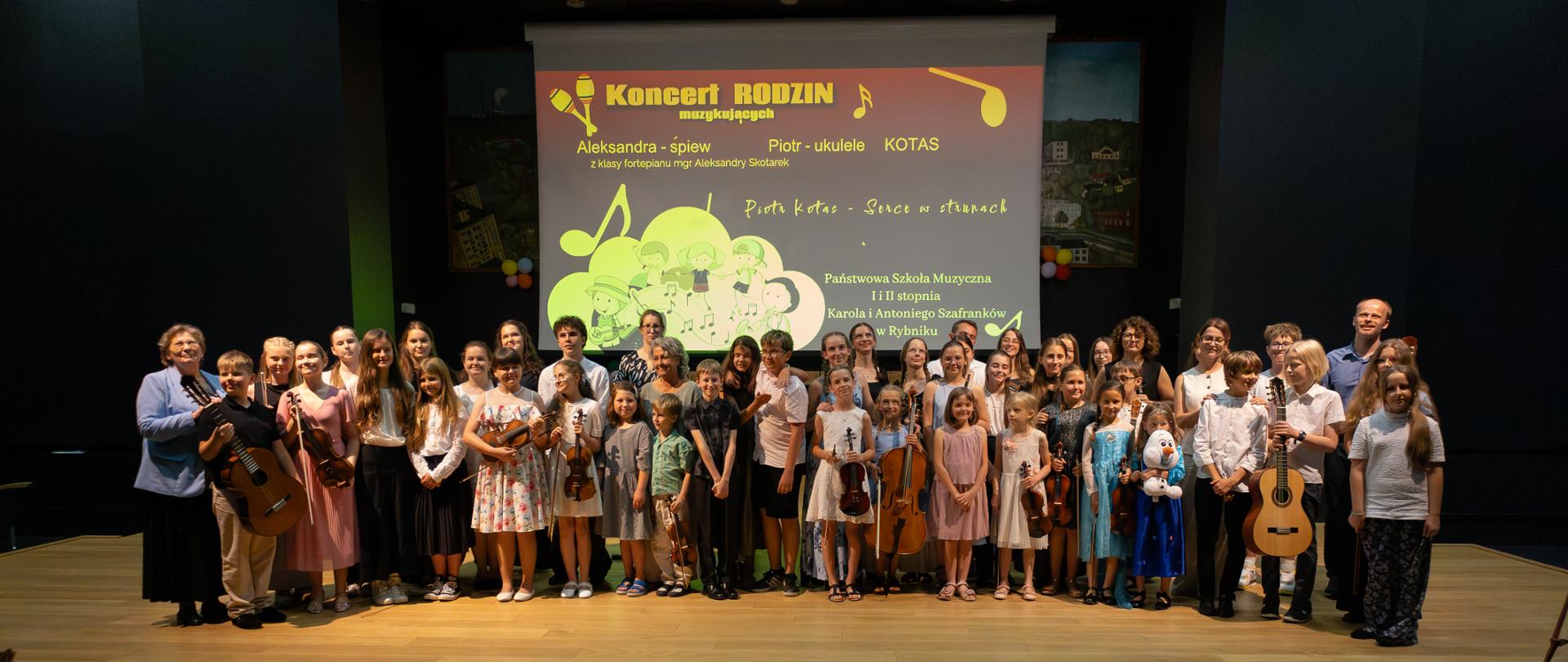 Duża grupa dzieci i młodzieży różnego wieku stoi na scenie w kilku rzędach po zakończonym występie. Młodzi artyści są ubrani w różnorodne, kolorowe stroje - od eleganckich sukienek, przez casualowe ubrania, po stroje folklorystyczne. Wśród nich znajdują się również dorośli opiekunowie i nauczyciele po bokach grupy.
