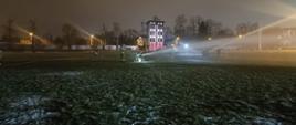 Nocne manewry taktyczne, na murawie stadionu ćwiczą zastępy strażaków, przy każdym zastępie widoczna doprowadzona linia wodna. w głębi po prawej widoczna wspinalnia