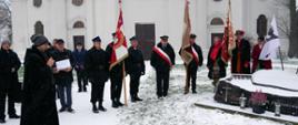 Kilkanaście osób zgromadzonych pod pomnikiem bł. ks. Jerzego Popiełuszki, cztery trzymają sztandary w dłoni, na pierwszym planie ksiądz czyta z książki tekst, w tle budynek bazyliki, pada śnieg