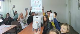 Na zdjęciu widać w budynku komendy powiatowej PSP w Ostrowcu Św. siedzącą młodzież z podniesionymi rękoma w których trzymają otrzymane czujki tlenku węgla oraz dymu. W tle prezentacja wyświetlana z rzutnika na temat zagrożeń związanych z czadem.