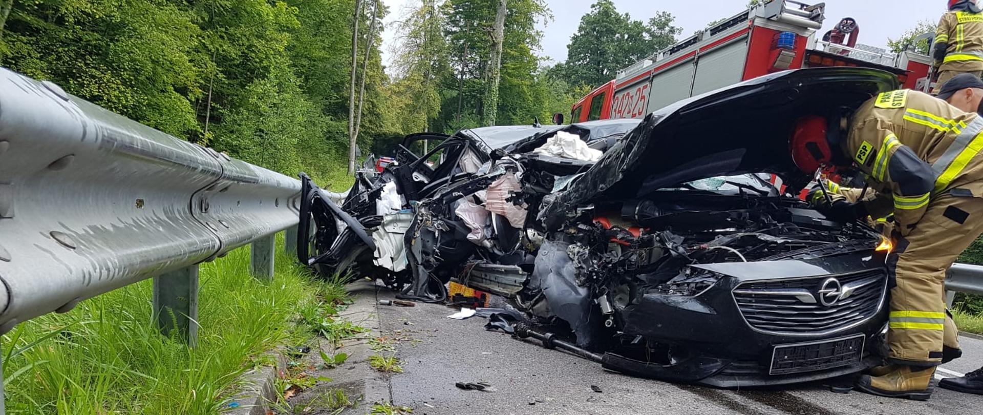 Rozbity na drodze wrak pojazdu, widoczne bardzo duże uszkodzenia prawej strony pojazdu. Za nim widoczny zarys pojazdu pożarniczego, po prawej stronie sylwetki strażaków.