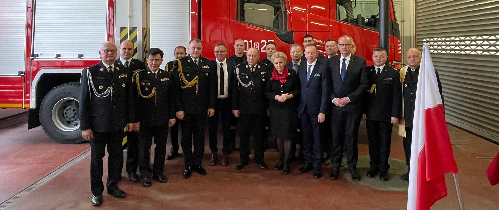 Fotografia wykonana w garażu jednostki ratowniczo-gaśniczej nr 1 komendy powiatowej Państwowej Straży Pożarnej w Mielcu. Grupowe zdjęcie zaproszonych gości i funkcjonariuszy. Funkcjonariusze ubrani w galowe mundury koloru granatowego Państwowej Straży Pożarnej. Zdjęcie wykonane na tle samochodu ratowniczo-gaśniczego przekazanego komendzie powiatowej Państwowej Straży Pożarnej w Mielcu.