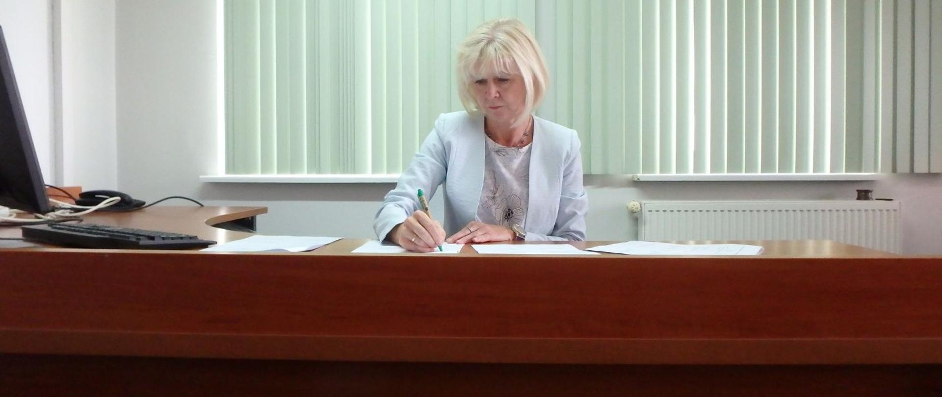 Regionalny Dyrektor Ochrony Środowiska w Olsztynie. Kobieta siedzi przy biurku, na którym leżą kartki. W ręku trzyma długopis.