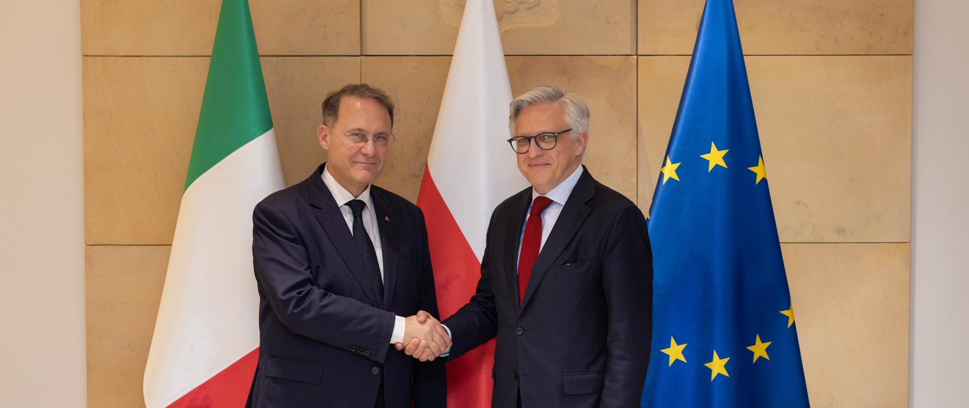 Dwóch mężczyzn wymienia uścisk dłoni na tle dużych flag: włoskiej, polskiej oraz UE: od prawej Podsekretarz Stanu Artur Harazim, od lewej: Wiceminister Spraw Zagranicznych i Współpracy Międzynarodowej Republiki Włoskiej Edmondo Cirielli 