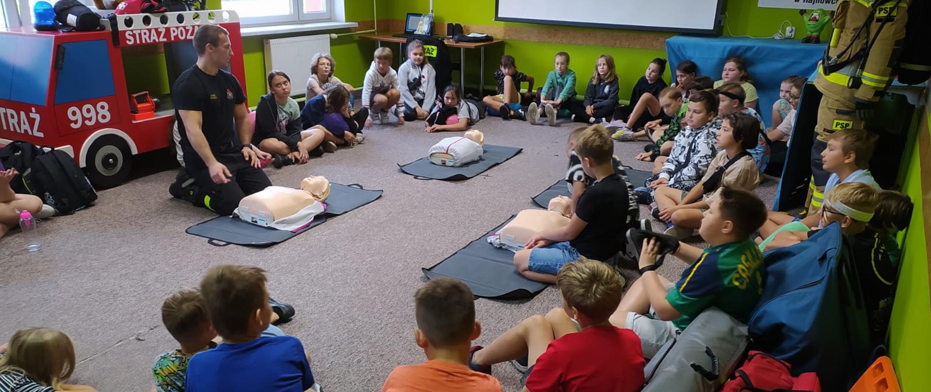 W pomieszczeniu na dywanie siedzi kilkadziesiąt dzieci obok siedzi strażak. Po środku leżą cztery manekiny.