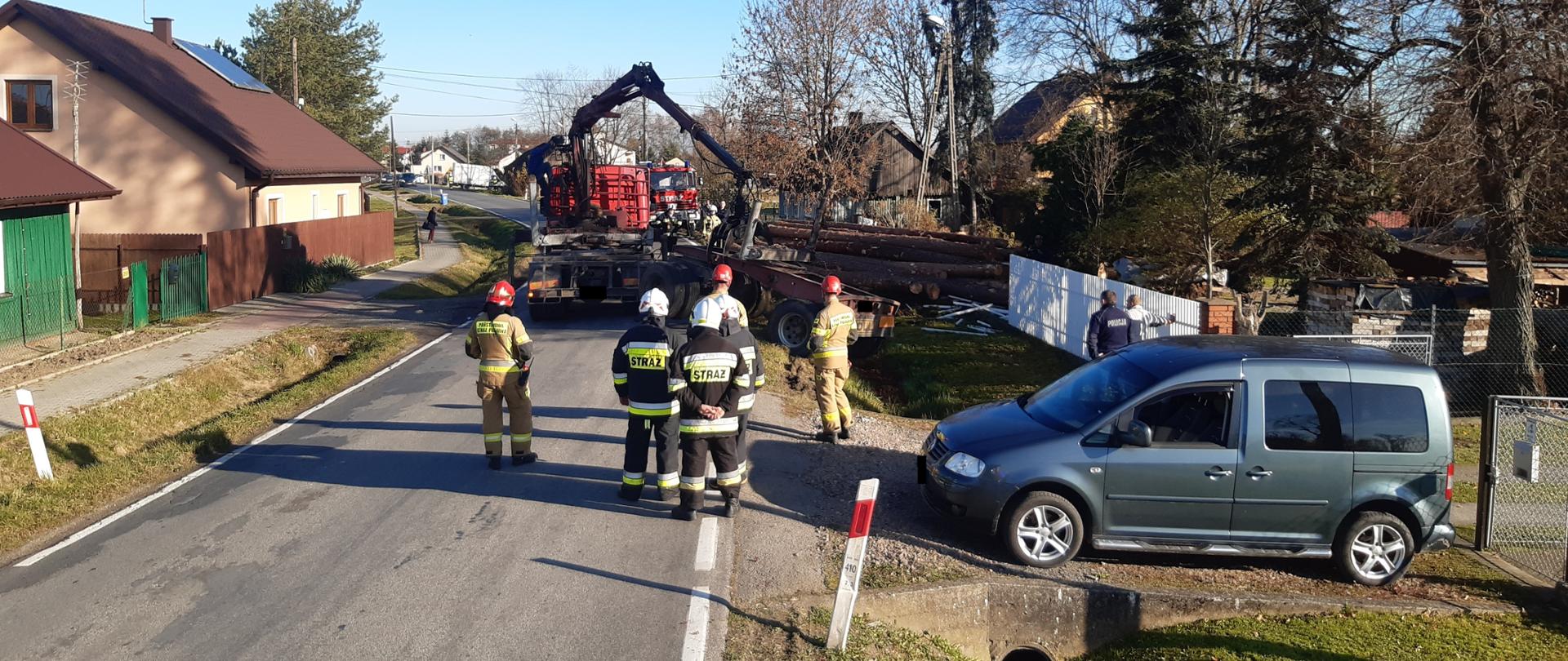 Zdjęcie przedstawia strażaków, samochód osobowy oraz naczepę do przewozu drzewa w rowie i samochód ciężarowy