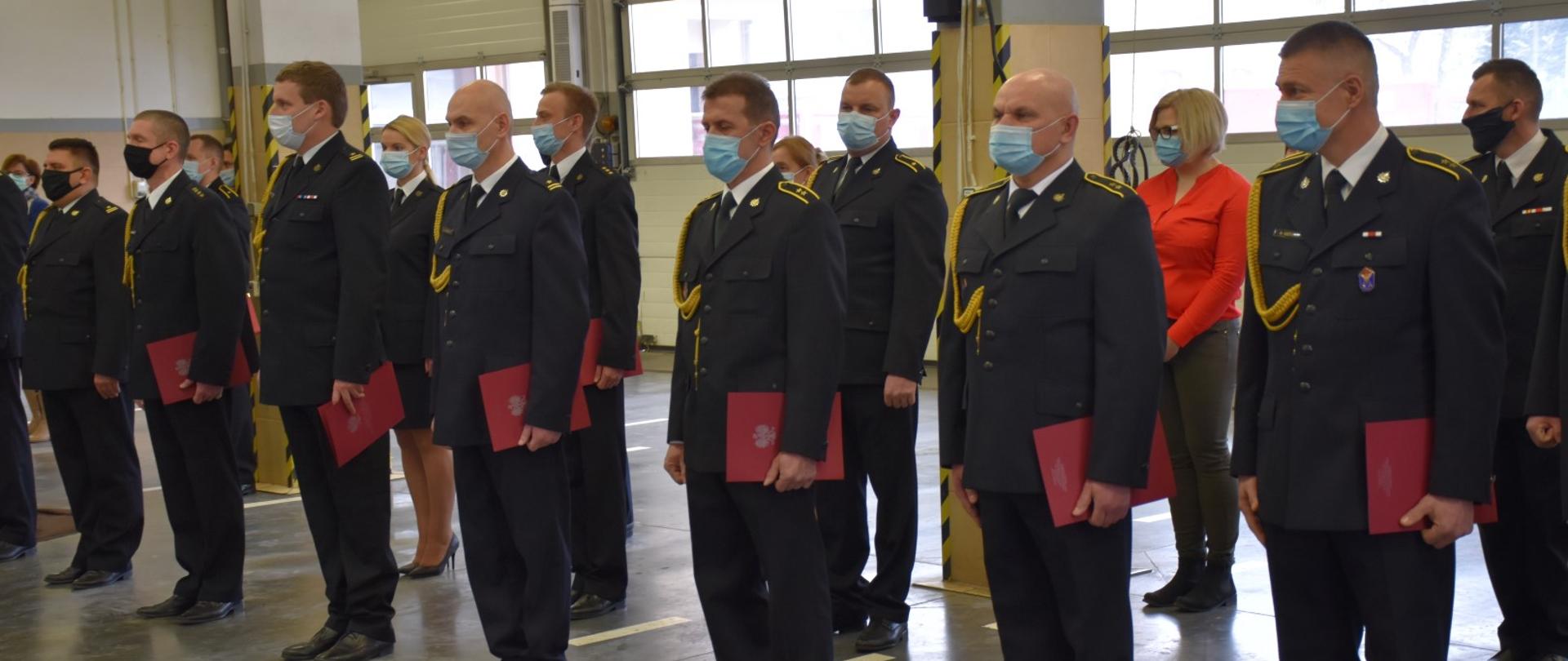 Zdjęcie przedstawia funkcjonariuszy Komendy Miejskiej PSP w Olsztynie stojących w pierwszym rzędzie z otrzymanymi decyzjami zwolnienia ze służby