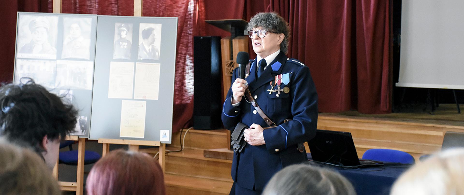 Krystyna Sobańska-Stępień w mundurze wojskowym z odznaczeniami przemawia do mikrofonu w sali z czerwonymi zasłonami (aula PLSP w Lublinie). W tle znajduje się tablica z historycznymi fotografiami i artykułami, fortepian oraz stolik z laptopem i mikrofonem.