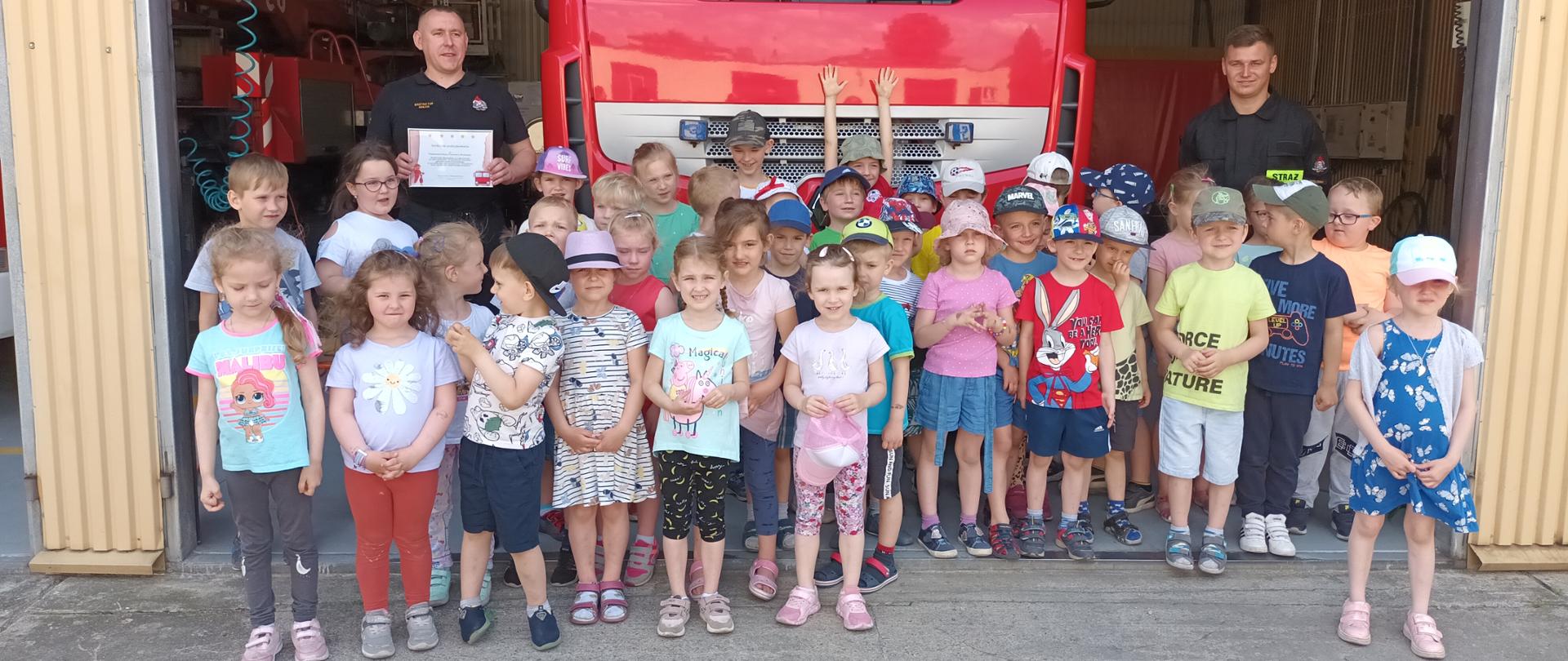 Zdjęcie przedstawia grupę przedszkolaków stojącą na tle dużego samochodu strażackiego. Auto stoi w garażu. Po bokach widoczni są strażacy, jeden z nich trzyma dyplom otrzymany od dzieci. W tle zdjęcia widoczny jest inny strażacki sprzęt.