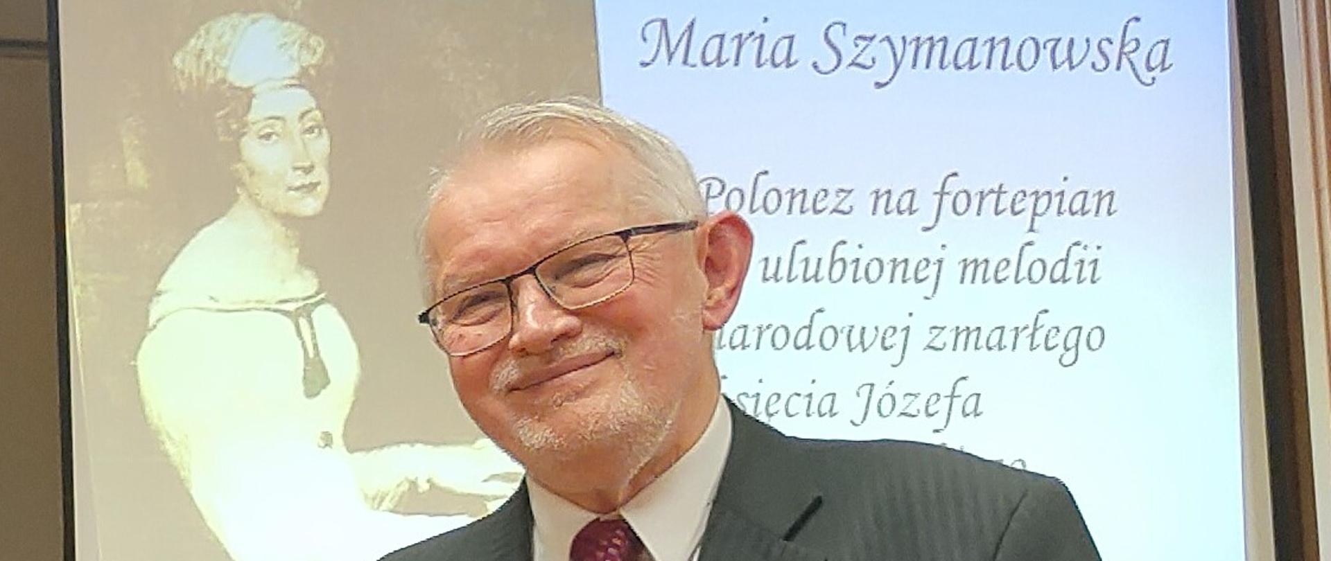 prof. Tadeusz Trzaskalik - stoi uśmiechnięty z bukietem pomarańczowych róż, w tle fragment prezentacji o Marii Szymanowskiej