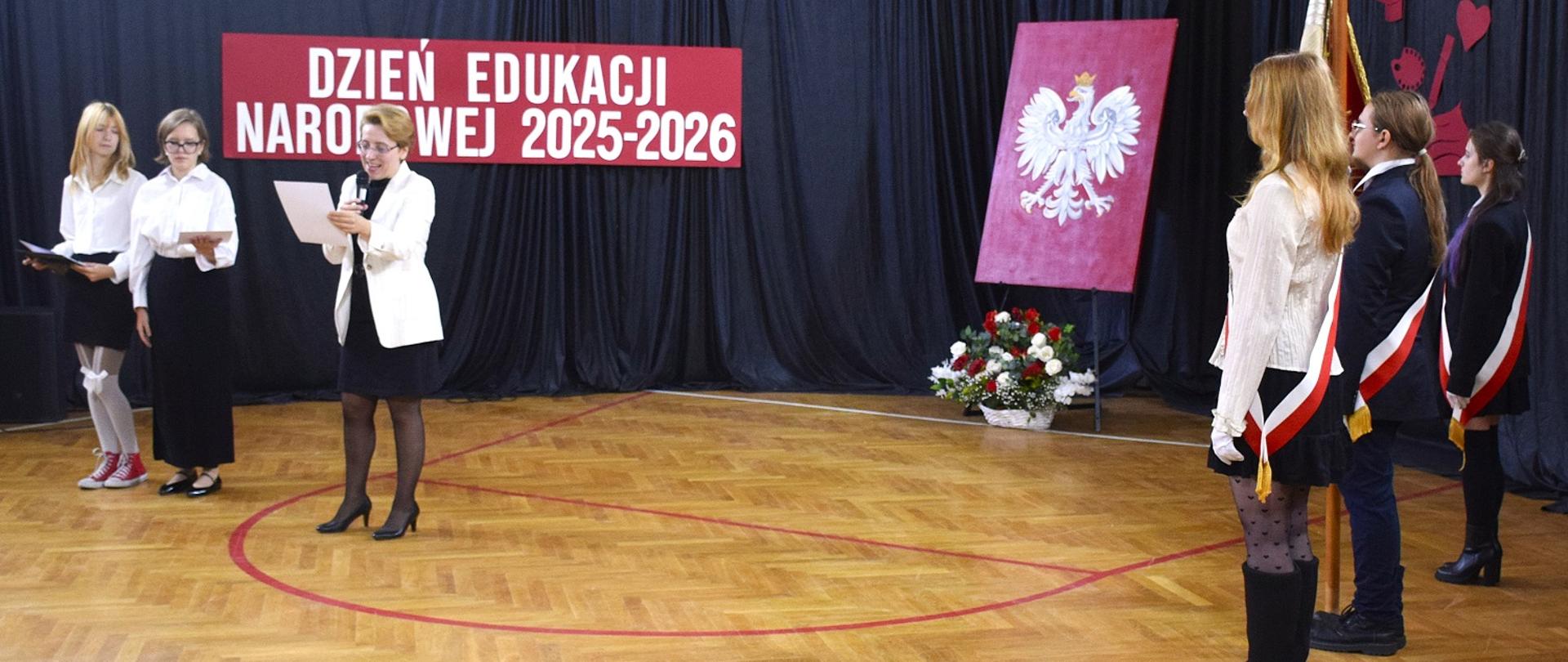 Na zdjęciu p. vice dyrektor, przedstawiciele samorządu uczniowskiego, poczet sztandarowy. W tle dekoracja: obraz - biały orzeł w koronie na czerwonym tle, biały napis: Dzień Edukacji Narodowej 2025-2026