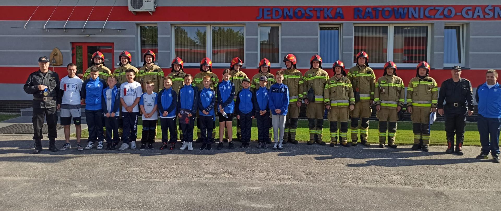 grupa młodych piłkarzy stoi przed grupa strażaków wszyscy się uśmiechają w tle widoczny budynek przed nimi szary asfalt.