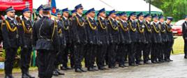 zdjęcie przedstawia pododdział strażaków podczas uroczystego dnia strażaka, z przodu stoi dowódca, pada rzęsisty deszcz