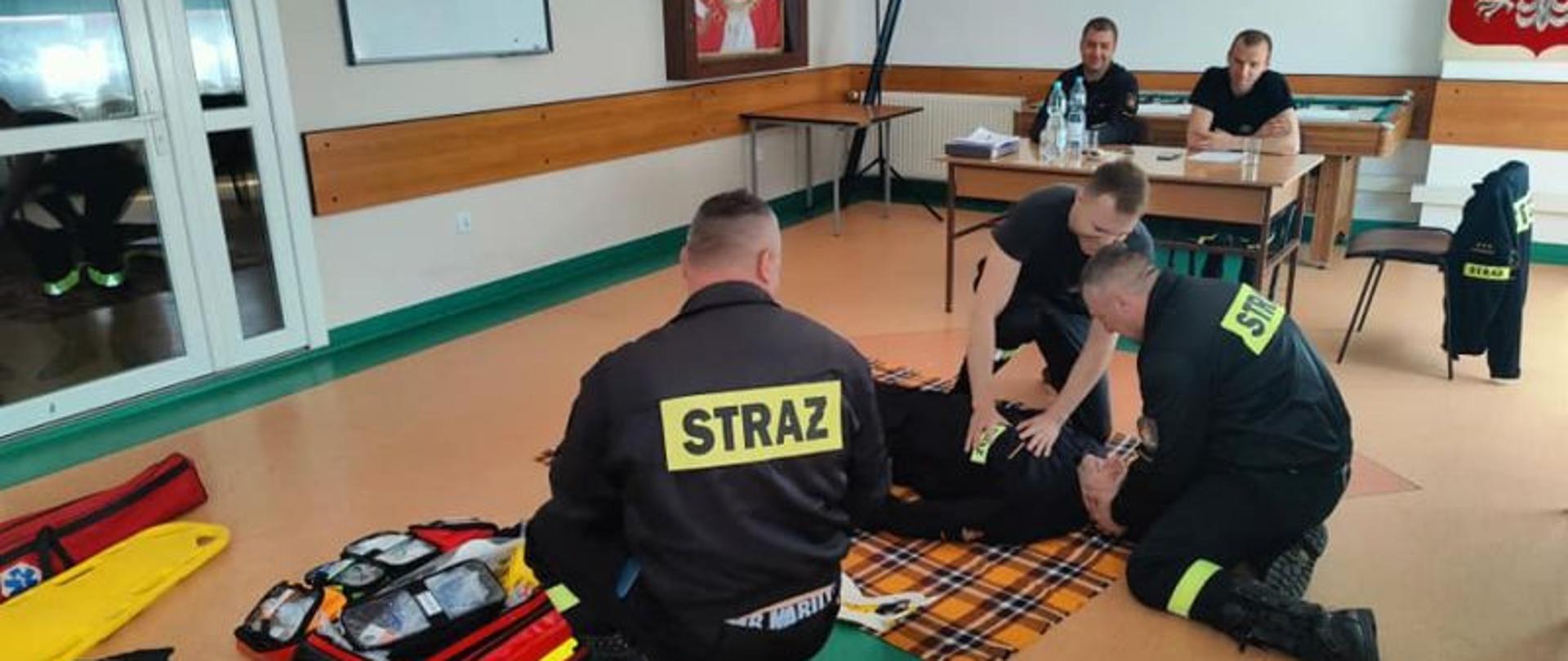 Zdjęcie wykonane na świetlicy przedstawia zaliczających scenkę strażaków. W tle komisja egzaminacyjną.