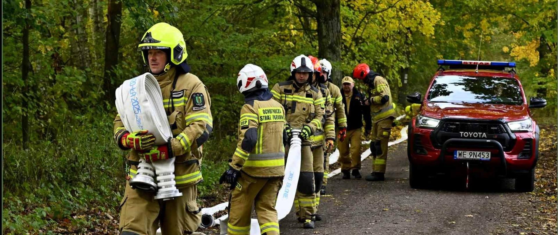 Na zdjęciu widać grupę strażaków w akcji, stojących na gruntowej drodze prowadzącej przez las, jesienią, co sugerują żółte i zielone liście na drzewach w tle.
W pierwszym planie, po lewej stronie, stoi strażak w pełnym umundurowaniu ochronnym w kolorze beżowo-żółtym i czerwonym hełmie (z elementami neonowo-żółtymi/zielonymi), trzymający zwinięte węże strażackie.
Za nim w rzędzie, ustawionym wzdłuż drogi, stoją kolejni strażacy w podobnym rynsztunku, niektórzy z białymi lub czerwonymi hełmami. Na kurtce jednego ze strażaków widoczny jest napis "OCHOTNICZA STRAŻ POŻARNA". Jeden z nich również trzyma wąż strażacki.
Po prawej stronie, obok rzędu strażaków, zaparkowany jest czerwony samochód terenowy, marki Toyota (widoczne logo), z napisem "STRAŻ" i niebieskimi światłami ostrzegawczymi na dachu. Tablica rejestracyjna samochodu to "WE 9N453".
Na ziemi widoczne są rozciągnięte węże strażackie.
