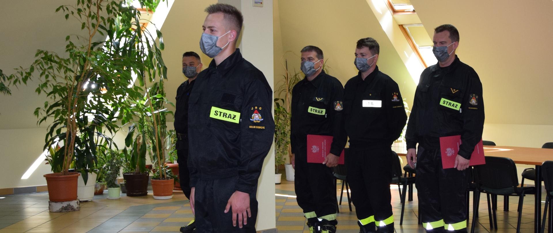 NA ZDJĘCIU NA PIERWSZYM PLANIE WIDAĆ STRAŻAKA W UBRANIU KOSZAROWYM, ZA NIM STOI 4 MĘŻCZYZN STRAŻAKÓW TAKŻE W UBRANIACH KOSZAROWYCH. WSZYSCY MĘŻCZYŹNI MAJĄ USTA I NOS ZABEZPIECZONE MASECZKAMI OCHRONNYMI.